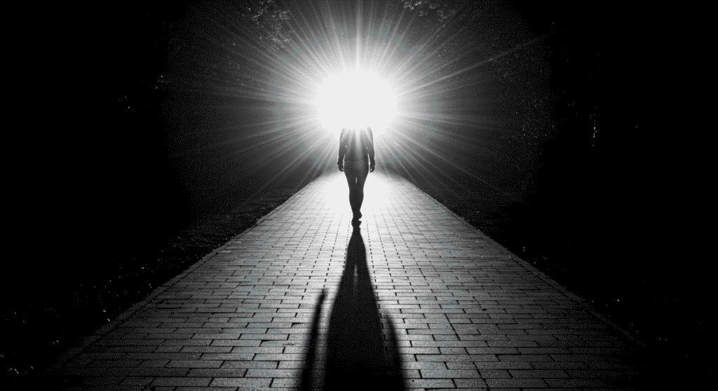 Fotografía en blanco y negro de una mujer caminando hacia una luz brillante al final de un camino. Imagen simbólica de esperanza y superación. Ideal para temas de inspiración, motivación y nuevos comienzos.