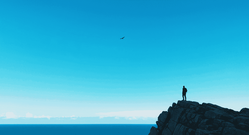 Paisaje costero con cielo azul, mar y una persona en la cima de una roca. Fotografía minimalista para transmitir calma, aventura y conexión con la naturaleza.

