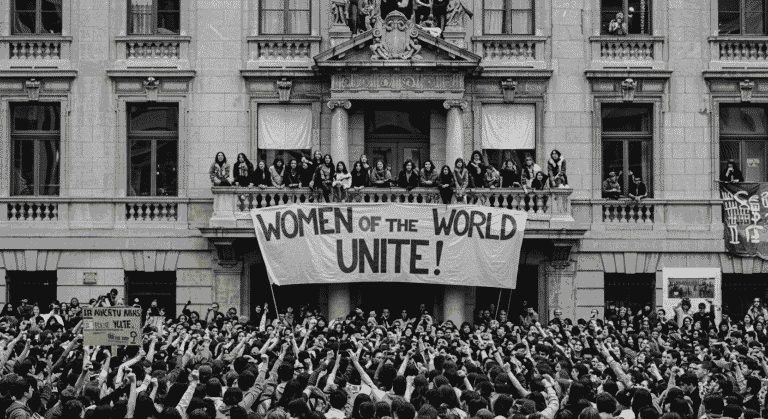"Fotografía en blanco y negro de una manifestación feminista. Imagen de una multitud protestando con un cartel que dice 'Mujeres del Mundo Uníos'. Ideal para contenido sobre historia, feminismo y movimientos sociales."