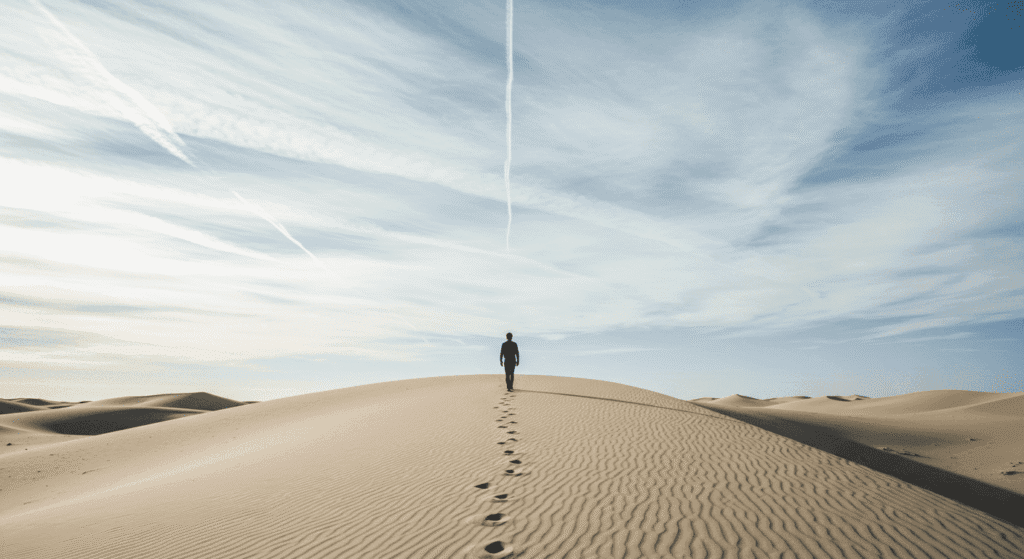 Persona caminando en duna de arena bajo cielo azul. Paisaje desértico con huellas en la arena. Imagen de viaje y aventura en el desierto.