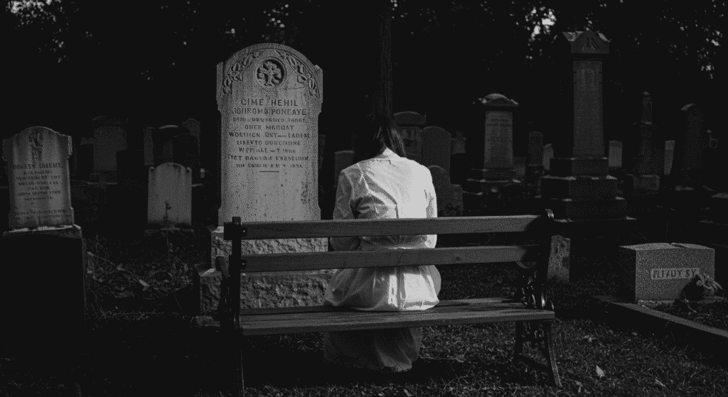 Fotografía en blanco y negro de persona sentada frente a lápida en cementerio de noche. Imagen sombría y melancólica con ambiente fantasmal. Luto y recuerdo.