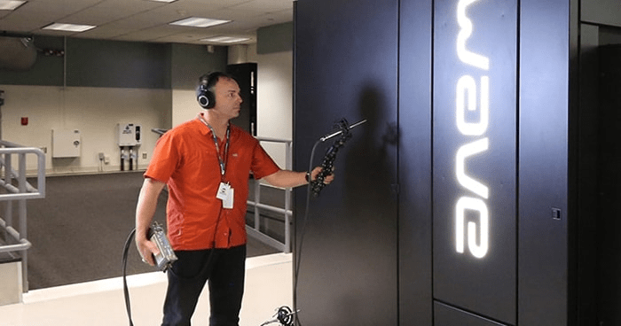 Computación Cuántica en Palabras Simples: Cómo Funciona y por Qué Importa 4 Un técnico con auriculares y camisa naranja examina un ordenador cuántico negro con la palabra "Wave" iluminada. Sostiene un dispositivo con una sonda. Fondo con una instalación moderna y limpia. Accesibilidad: imagen que muestra el trabajo de un técnico con tecnología de computación cuántica