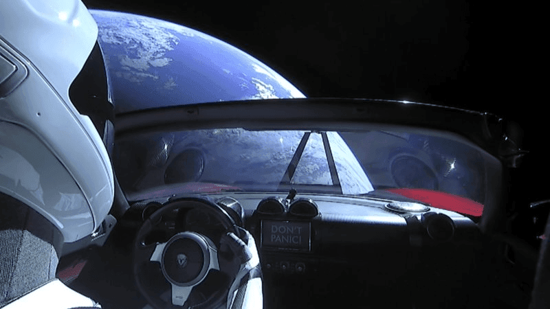 SpaceX y Falcon Heavy: Cómo Revoluciona la Exploración Espacial 4 Vista interior de un Tesla Roadster rojo en el espacio. Un maniquí con traje espacial conduce, con la Tierra visible a través del parabrisas. La pantalla del coche muestra 'DON'T PANIC!'. Accesibilidad: imagen que muestra un coche Tesla en el espacio