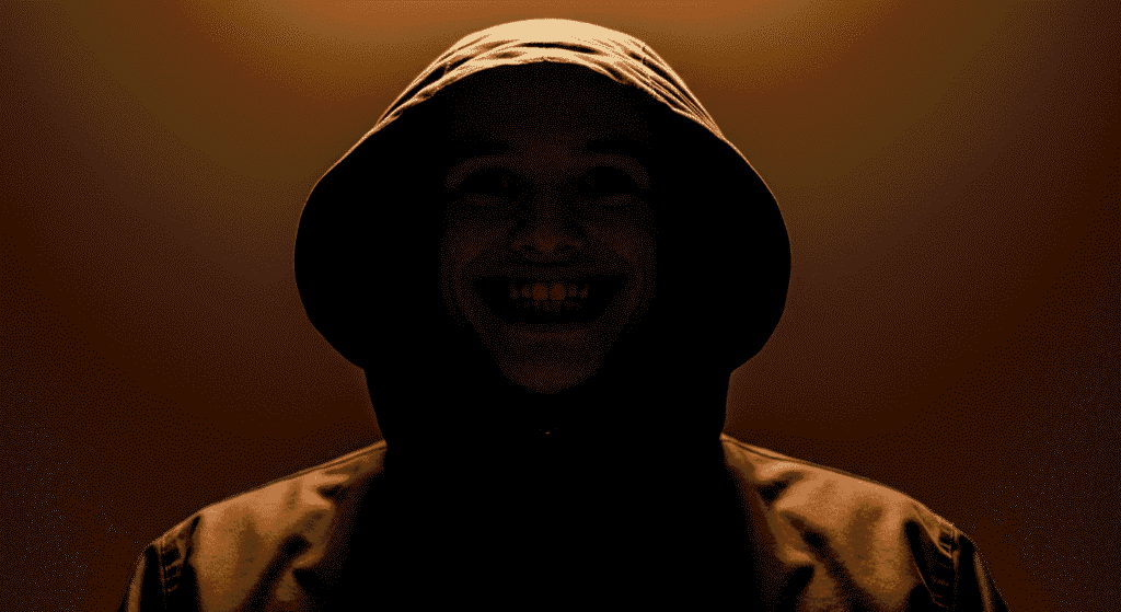 El crimen de Gádor: la escalofriante historia del verdadero hombre del saco 6 Primer plano de rostro siniestro con sonrisa malvada bajo capucha en ambiente cálido. Fotografía de terror psicológico con iluminación dramática.