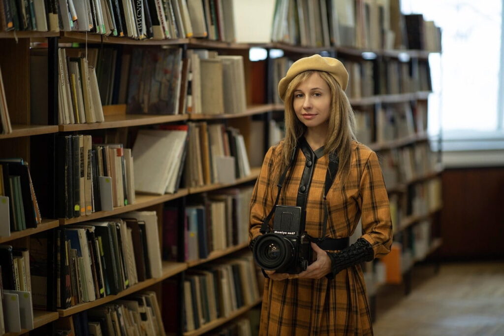 La operación secreta detrás de Argo: La historia real que inspiró el cine 4 Una mujer en una biblioteca rodeada de estantes llenos de libros, vestida con un atuendo retro y sosteniendo una cámara fotográfica profesional.