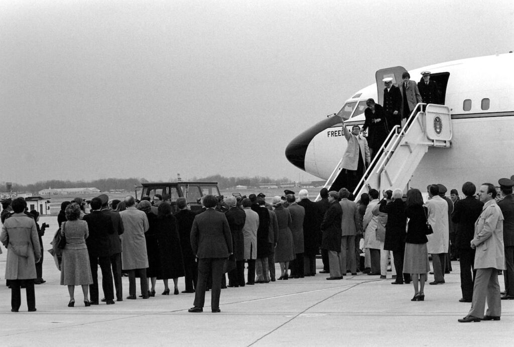 La operación secreta detrás de Argo: La historia real que inspiró el cine 6 Fotografía en blanco y negro mostrando un grupo de personas reunidas en una pista de aterrizaje observando a varios pasajeros descender por la escalera de un avión con la inscripción ‘Freedom’ en el costado.
