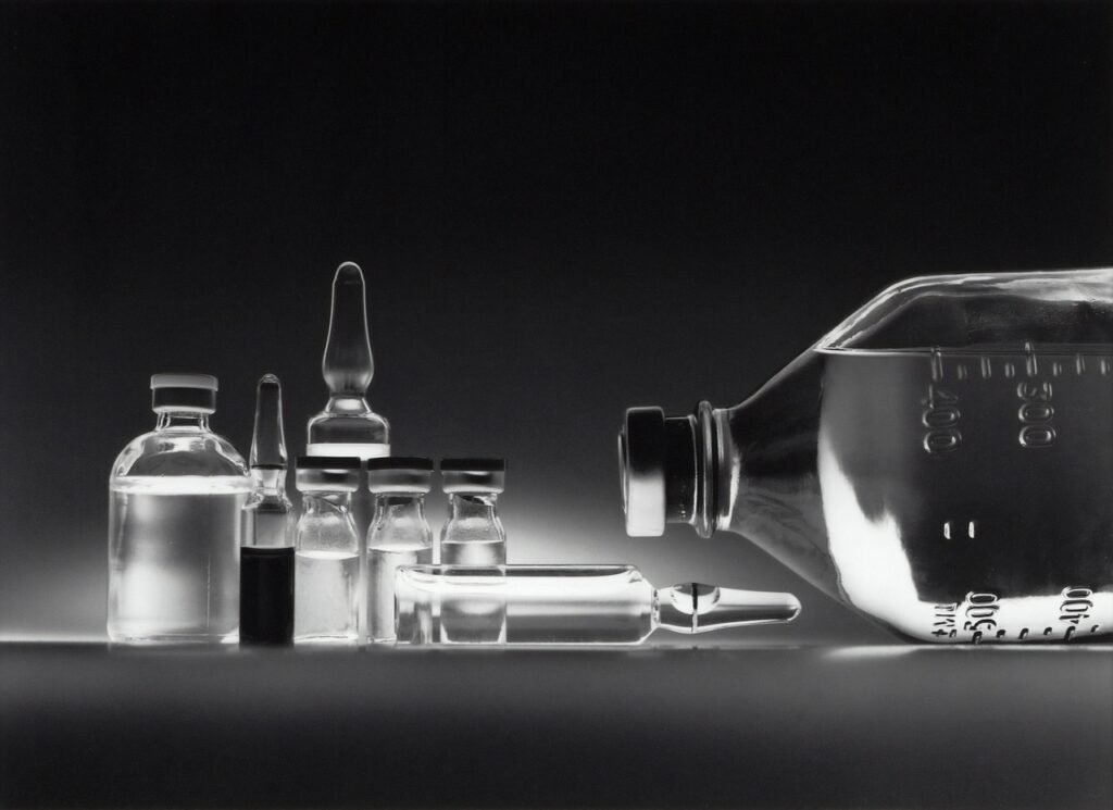 HMPV: Innovaciones en el Control de Infecciones Respiratorias 6 Un conjunto de viales de vidrio transparente y una botella médica grande colocados sobre una superficie oscura, iluminados de manera que resaltan los detalles del vidrio y las mediciones. La imagen evoca un entorno clínico o de laboratorio enfocado en investigación o almacenamiento de medicamentos.