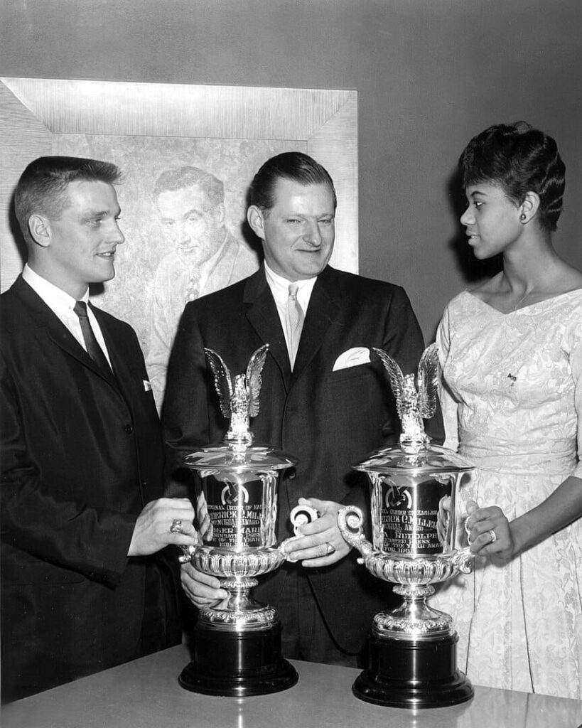 Historias inspiradoras: Atletas que convirtieron los obstáculos en triunfos 1 Tres personas sosteniendo trofeos, con un fondo enmarcado de una pintura. La mujer a la derecha luce elegante y los hombres visten trajes formales, celebrando un momento importante.