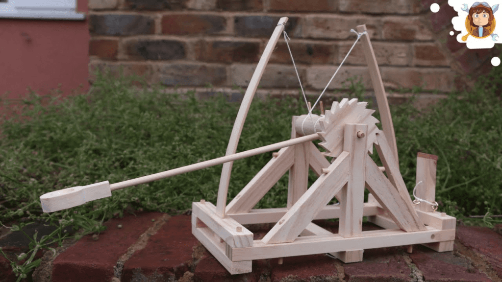 Inventos de Leonardo da Vinci: Parte 2 6 Modelo de trabuco o catapulta antigua. Recreación en madera de máquina de asedio medieval. Diseño con brazo de palanca, rueda dentada y estructura simple. Maqueta educativa sobre base de ladrillos rojos