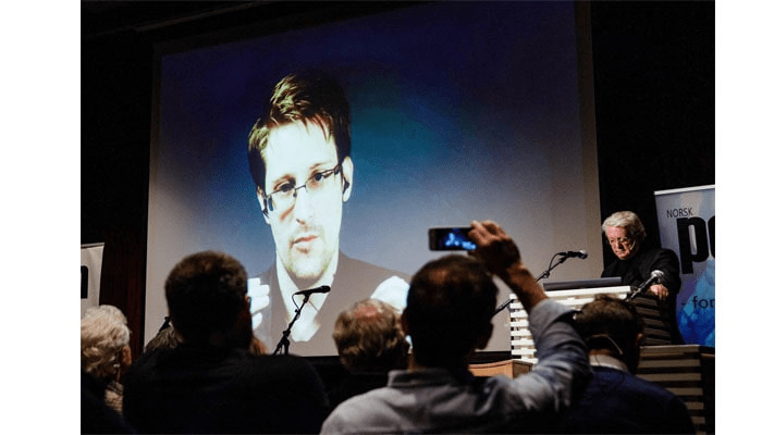 El caso Edward Snowden 5 Edward Snowden en presentación remota a través de pantalla grande. Audiencia viendo conferencia con Snowden virtualmente. Persona grabando con teléfono móvil la presentación. Evento tecnológico y participación virtual