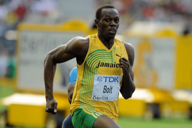 Usain Bolt corriendo en carrera de atletismo. El atleta jamaiquino lleva su uniforme deportivo y está en plena competición.