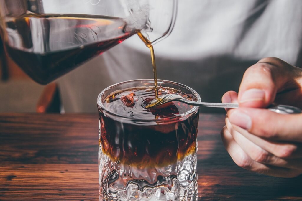 6 Cosas que Probablemente No Sabías sobre el Café (Pero Deberías Saber) 5 Mano vertiendo café recién preparado desde una jarra de vidrio en un vaso con hielo, con iluminación cálida y acogedora.