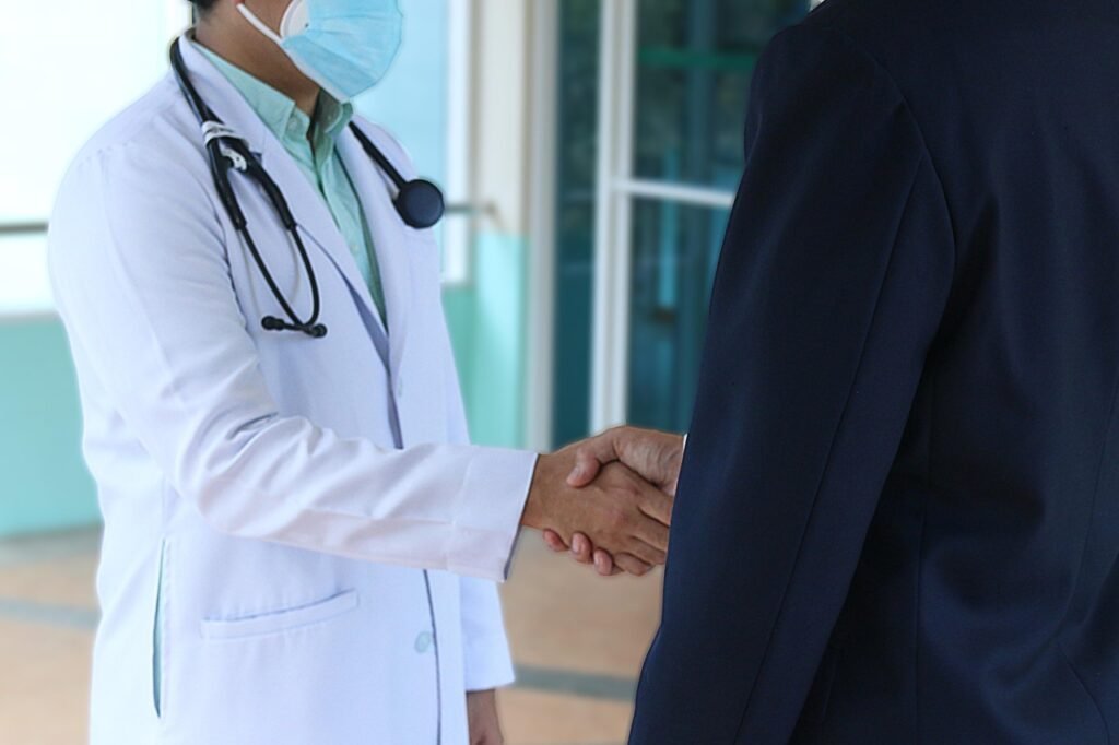 Trastornos de la Conducta Alimentaria: Lo Que Nadie Te Cuenta 6 Doctor con bata blanca, mascarilla y estetoscopio estrechando la mano de una persona con traje formal en un entorno moderno y luminoso.