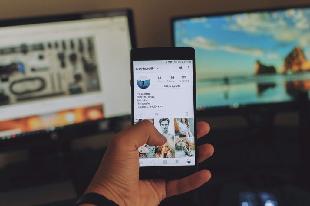 El Peligro de la Falsa Autoestima: Cómo Detectarla y Fortalecer tu Confianza 6 Mano sosteniendo un smartphone con un perfil de Instagram en pantalla, frente a dos monitores de computadora en un espacio de trabajo.