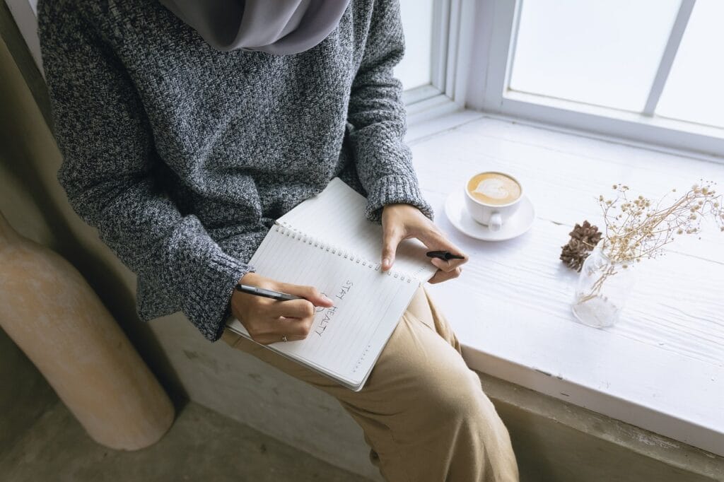 El Peligro de la Falsa Autoestima: Cómo Detectarla y Fortalecer tu Confianza 2 Persona escribiendo "Stay Healthy" en un cuaderno junto a una ventana iluminada, con una taza de café y flores secas sobre el alféizar.