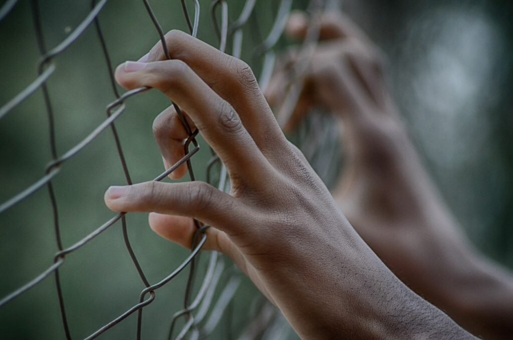 Pena de Muerte: Orígenes, Métodos y su Impacto en la Sociedad 6 Primer plano de manos humanas sujetando con fuerza una cerca de alambre, con fondo verde borroso. Representación de cautiverio y deseo de libertad.