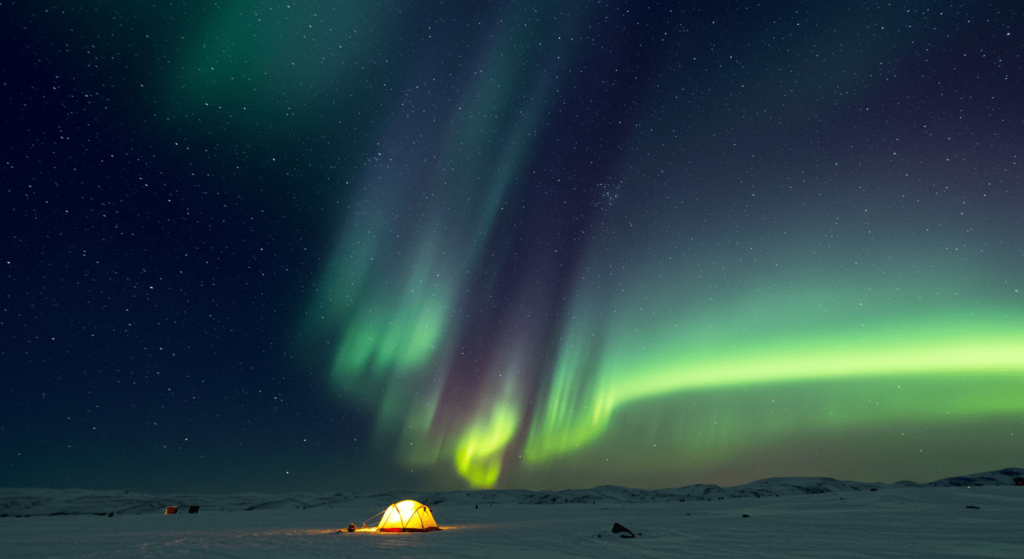 ¿Qué son las auroras polares? El misterio y la ciencia detrás de las luces del norte y sur 3 Aurora boreal multicolor sobre paisaje nevado con una tienda de campaña iluminada. Aventura y maravilla en el Ártico. Accesible para personas con discapacidad visual.