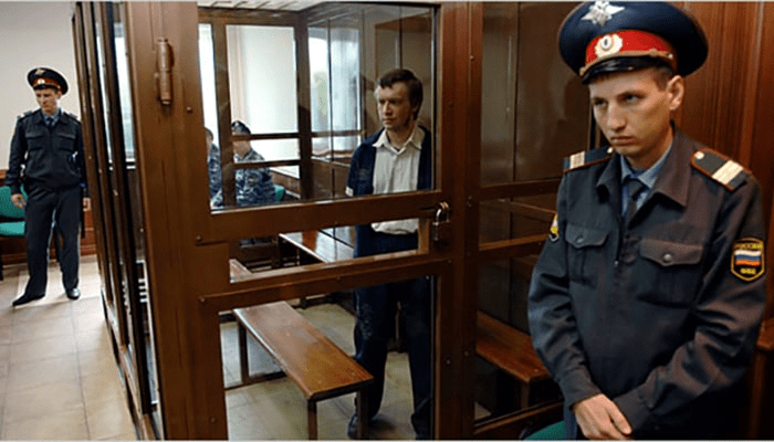 Alexander Pichushkin, el “Maníaco del Martillo”: La historia del asesino del ajedrez 5 Imagen de sala de juicio con acusado y policías. Fotografía de tribunal con jaula de acusado y agentes uniformados. Escena judicial con detenido y fuerzas de seguridad.