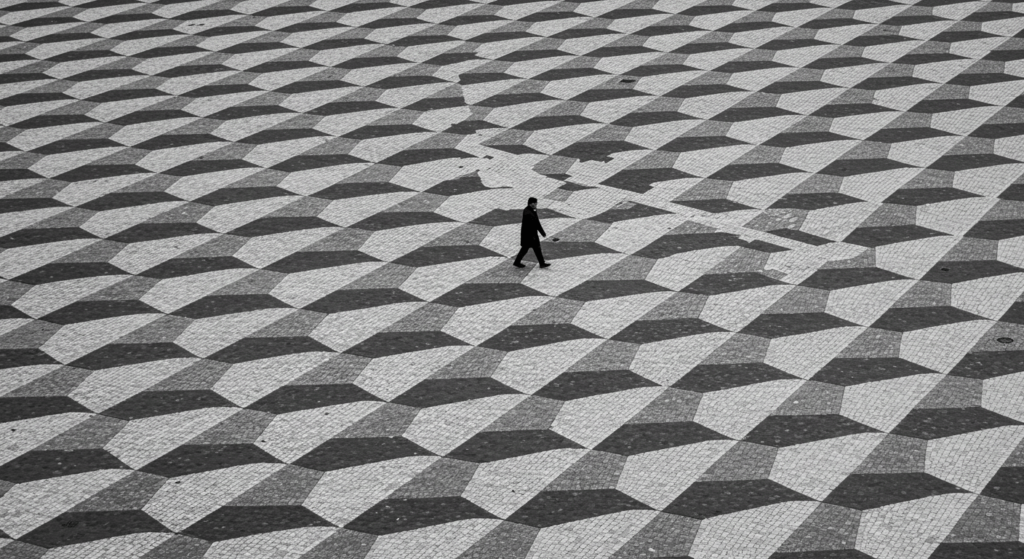 ¿Qué es el minimalismo? La filosofía de vivir con lo esencial 6 "Plaza geométrica: fotografía en blanco y negro de un hombre caminando sobre un suelo con diseño de triángulos. Ideal para contenido sobre arquitectura urbana, soledad y diseño. Accesible para personas con discapacidad visual."