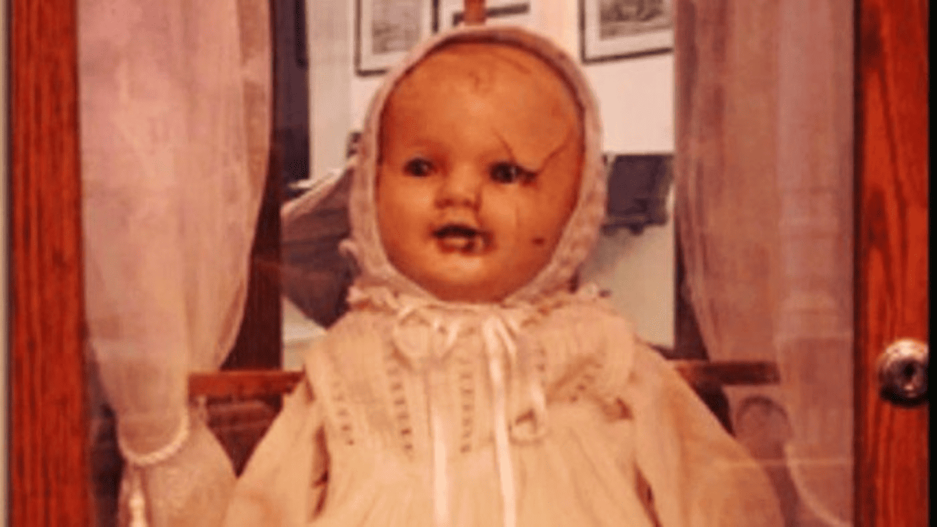Muñecos poseídos: 5 historias reales tan aterradoras que parecen ficción 5 Muñeca antigua con grietas y vestido blanco. Juguete vintage dañado en interior con cortinas. Imagen para accesibilidad y posicionamiento SEO.