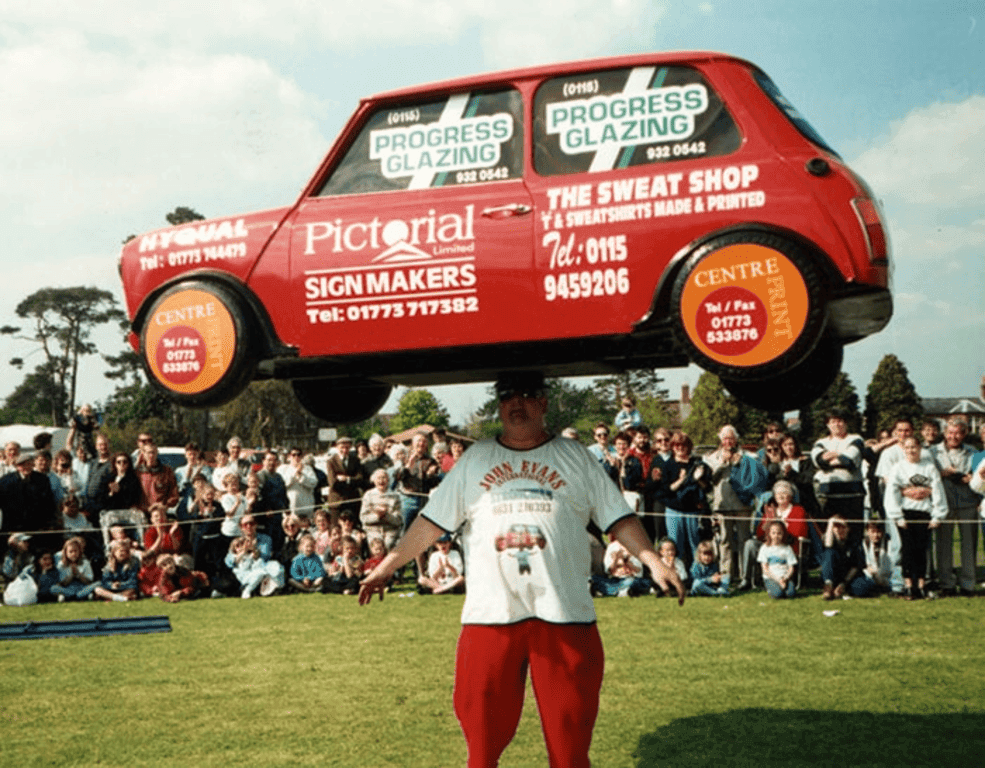 6 Récords Guinness increíbles que te dejarán con la boca abierta 2 Hombre fuerte levanta un Mini Cooper rojo lleno de publicidad. John Evans mostrando su fuerza ante una multitud. Récord de levantamiento de peso.