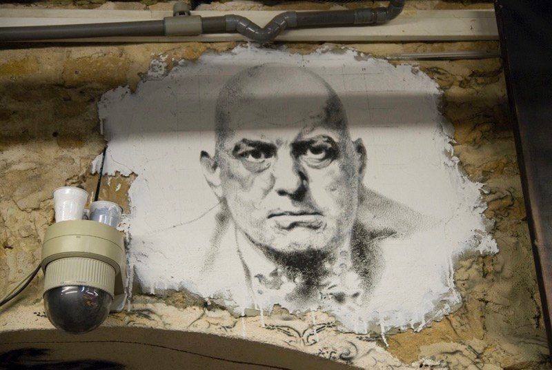 Aleister Crowley: el enigma del “Bestia 666” y los mitos que lo convirtieron en leyenda 1 Retrato mural en blanco y negro de Aleister Crowley pintado sobre una pared de piedra, con una expresión seria y una cámara de seguridad al costado.